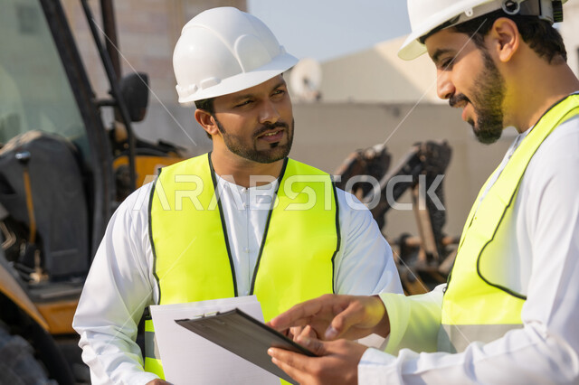 تبادل المعلومات والخبرة، دراسة المخطط والمشروع على ارض الواقع، وظيفة هندسية شبابية، مهندسان عربيان خليجيان اماراتيان يرتديان الكندورة والسترة وخوذة الحماية يعملان في القطاع الهندسي في الامارات، مراقبة ومتابعة عمليات البناء، مفهوم ادارة المشاريع الهندسية والانشاءات