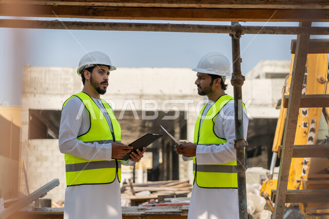 تبادل المعلومات والخبرة، دراسة المخطط والمشروع على ارض الواقع، وظيفة هندسية شبابية، مهندسان عربيان خليجيان اماراتيان يرتديان الكندورة والسترة وخوذة الحماية يعملان في القطاع الهندسي في الامارات، مراقبة ومتابعة عمليات البناء، مفهوم ادارة المشاريع الهندسية والانشاءات