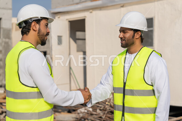 إيماءات المصافحة والترحيب والاستقبال, مفهوم التعاون والعمل المشترك, مهندسان عربيان خليجيان اماراتيان يرتديان الخوذة وسترة الحماية يقومان بالتصافح والاتفاق على الخطط والمشاريع الهندسية في موقع العمل, الإنشاءات المعمارية في الأراضي الاماراتية