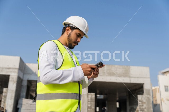 استخدام جهاز تقني تكنولوجي، مهنة هندسية شبابية، مهندس عربي خليجي اماراتي يرتدي الكندورة والسترة والخوذة يمسك الهاتف المحمول، العمل في القطاع الهندسي في الامارات، الاشرف على عمليات البناء، ادارة المشاريع الهندسية والانشاءات، دراسة المخطط والمشروع على ارض الواقع