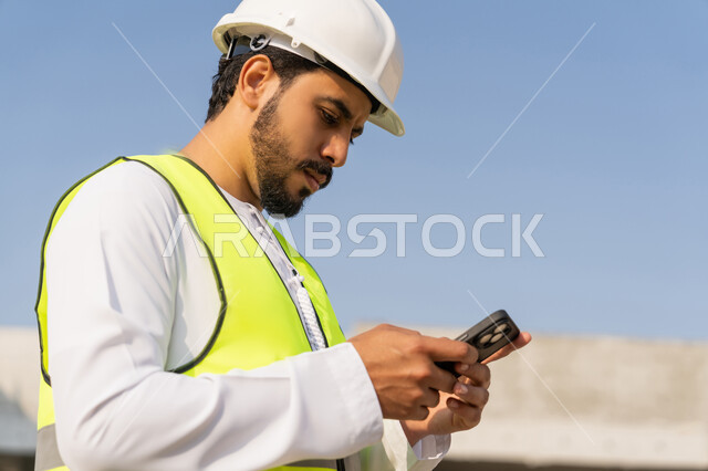Using a technological device, a youth engineering profession, an Arab Gulf Emirati engineer wearing a kandura, jacket and helmet holding a mobile phone, working in the engineering sector in the Emirates, supervising construction operations, managing engineering and construction projects, studying the plan and the project on the ground