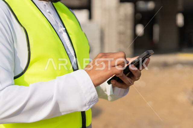 Using a technological device, a youth engineering profession, an Arab Gulf Emirati engineer wearing a kandura, jacket and helmet holding a mobile phone, working in the engineering sector in the Emirates, supervising construction operations, managing engineering and construction projects, studying the plan and the project on the ground