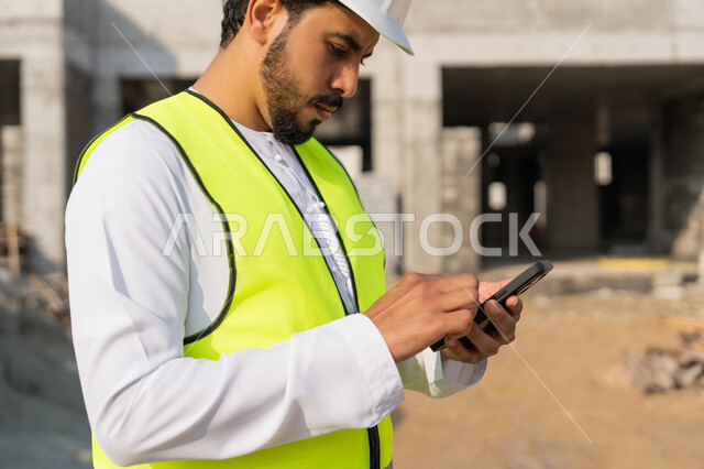 Using a technological device, a youth engineering profession, an Arab Gulf Emirati engineer wearing a kandura, jacket and helmet holding a mobile phone, working in the engineering sector in the Emirates, supervising construction operations, managing engineering and construction projects, studying the plan and the project on the ground