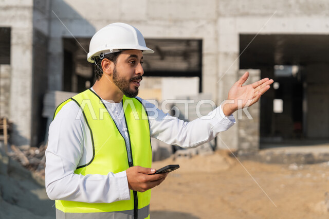 Using a technological device, a youth engineering profession, an Arab Gulf Emirati engineer wearing a kandura, jacket and helmet holding a mobile phone, working in the engineering sector in the Emirates, supervising construction operations, managing engineering and construction projects, studying the plan and the project on the ground