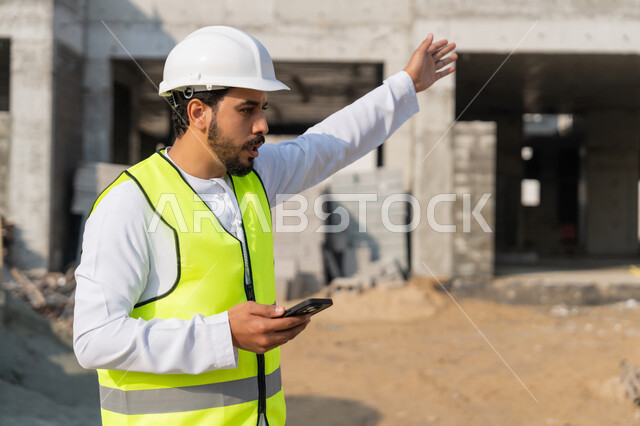 Using a technological device, a youth engineering profession, an Arab Gulf Emirati engineer wearing a kandura, jacket and helmet holding a mobile phone, working in the engineering sector in the Emirates, supervising construction operations, managing engineering and construction projects, studying the plan and the project on the ground