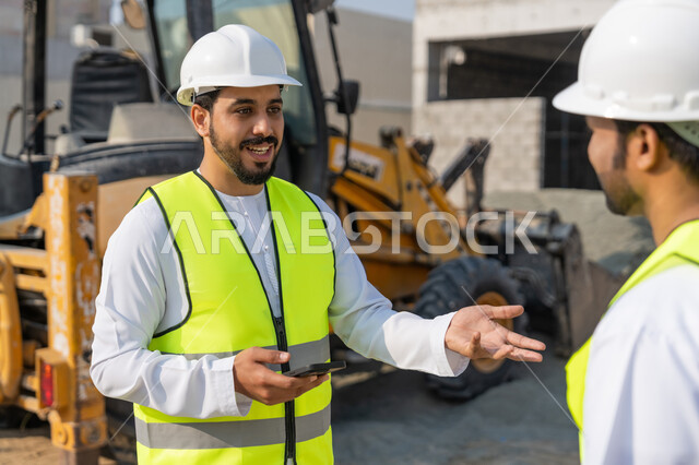 Using a technological device, a youth engineering profession, an Arab Gulf Emirati engineer wearing a kandura, jacket and helmet holding a mobile phone, working in the engineering sector in the Emirates, supervising construction operations, managing engineering and construction projects, studying the plan and the project on the ground