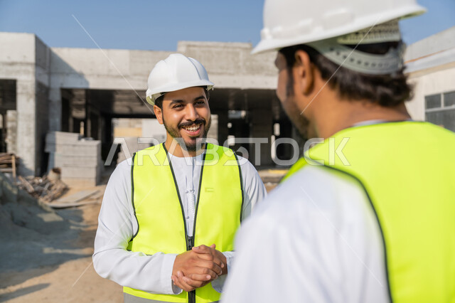 Exchange of information and experience, study of the plan and project on the ground, a youth engineering job, two Arab Gulf Emirati engineers wearing kandura, jacket and protective helmet working in the engineering sector in the Emirates, monitoring and following up on construction operations, the concept of engineering and construction project management.