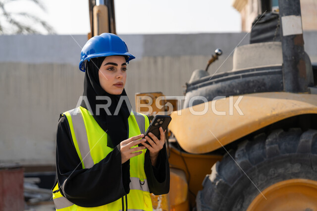 استخدام جهاز تقني حديث، مهنة ووظيفة نسائية، إمرأة عربية خليجية سعودية ترتدي السترة والخوذة تقوم بالاشراف على مبنى قيد الإنشاء والتشييد، مفهوم الهندسة الانشائية والمعمارية، مهندسة اماراتية تمسك بيدها الهاتف المحمول، أعمال البناء وصب الخرسانات ووضع الأساسات الأولية