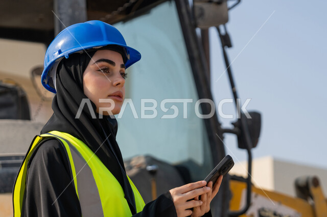استخدام جهاز تقني حديث، مهنة ووظيفة نسائية، إمرأة عربية خليجية سعودية ترتدي السترة والخوذة تقوم بالاشراف على مبنى قيد الإنشاء والتشييد، مفهوم الهندسة الانشائية والمعمارية، مهندسة اماراتية تمسك بيدها الهاتف المحمول، أعمال البناء وصب الخرسانات ووضع الأساسات الأولية
