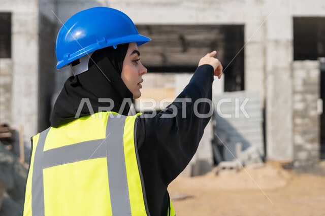 الاشارة الى شيء ما، إمرأة اماراتية تشرف على أعمال البناء وصب الخرسانات ووضع الأساسات الأولية، مهنة ووظيفة نسائية، صورة مقربة لمهندسة عربية خليجية سعودية محجبة ترتدي السترة والخوذة تقوم بمتابعة مبنى قيد الإنشاء والتشييد, مفهوم الهندسة الانشائية والمعمارية