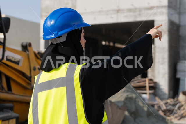 الاشارة الى شيء ما، إمرأة اماراتية تشرف على أعمال البناء وصب الخرسانات ووضع الأساسات الأولية، مهنة ووظيفة نسائية، صورة مقربة لمهندسة عربية خليجية سعودية محجبة ترتدي السترة والخوذة تقوم بمتابعة مبنى قيد الإنشاء والتشييد, مفهوم الهندسة الانشائية والمعمارية