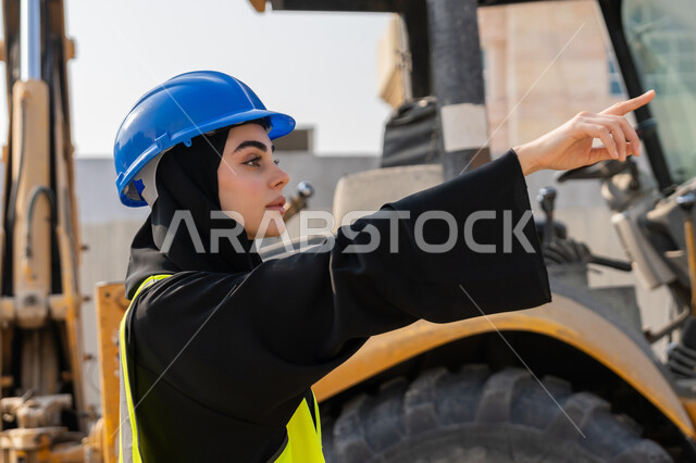 الاشارة الى شيء ما، إمرأة اماراتية تشرف على أعمال البناء وصب الخرسانات ووضع الأساسات الأولية، مهنة ووظيفة نسائية، صورة مقربة لمهندسة عربية خليجية سعودية محجبة ترتدي السترة والخوذة تقوم بمتابعة مبنى قيد الإنشاء والتشييد, مفهوم الهندسة الانشائية والمعمارية