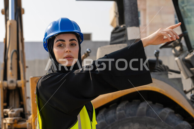 الاشارة الى شيء ما، إمرأة اماراتية تشرف على أعمال البناء وصب الخرسانات ووضع الأساسات الأولية، مهنة ووظيفة نسائية، صورة مقربة لمهندسة عربية خليجية سعودية محجبة ترتدي السترة والخوذة تقوم بمتابعة مبنى قيد الإنشاء والتشييد, مفهوم الهندسة الانشائية والمعمارية