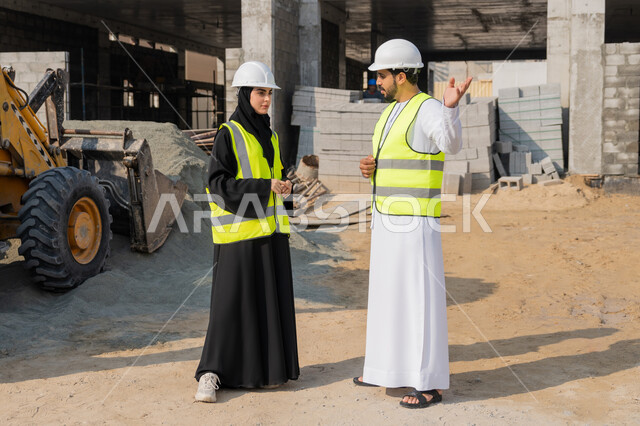 Engineering and construction project management, studying the plan and the project on the ground, an engineering profession and job, an Arab Gulf Emirati engineer wearing a kandura, jacket and protective helmet working with his colleague in the engineering sector in the United Arab Emirates, monitoring and following up on construction operations.