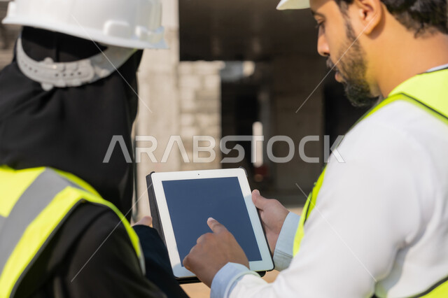 استخدام جهاز التابلت التقني في المجال الهندسي، دراسة المخطط على ارض الواقع، وظيفة ومهنة هندسية، مهندس عربي خليجي اماراتي يرتدي الكندورة والسترة وخوذة الحماية يعمل مع زميلته في القطاع الهندسي في الامارات، ادارة المشاريع الهندسية والانشاءات باستخدام التكنولوجيا