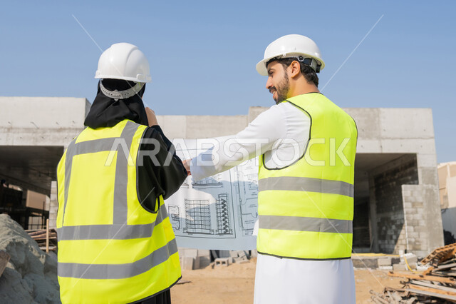 Follow-up and supervision of engineering and construction projects, studying the plan and the project on the ground, an engineering profession and job, an Arab Gulf Emirati engineer wearing a kandura, jacket and protective helmet working with his colleague in the engineering sector in the United Arab Emirates, monitoring and managing construction operations.