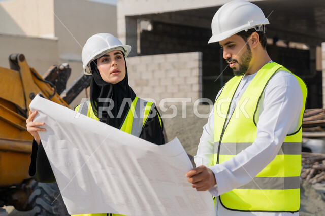 Engineering and construction project management, studying the plan and the project on the ground, an engineering profession and job, an Arab Gulf Emirati engineer wearing a kandura, jacket and protective helmet working with his colleague in the engineering sector in the United Arab Emirates, monitoring and following up on construction operations.