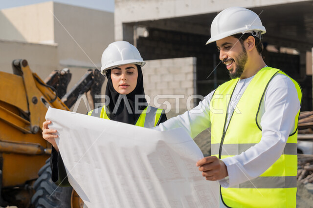 المتابعة والاشراف على المشاريع الهندسية والانشاءات، دراسة المخطط والمشروع على ارض الواقع، مهنة ووظيفة هندسية، مهندس عربي خليجي اماراتي يرتدي الكندورة والسترة وخوذة الحماية يعمل مع زميلته في القطاع الهندسي في الامارات العربية المتحدة، مراقبة وادارة عمليات البناء