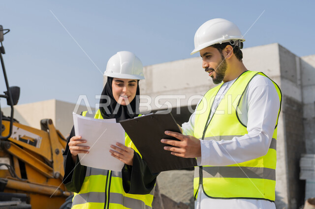 ادارة المشاريع الهندسية والانشاءات، دراسة المخطط والمشروع على ارض الواقع، مهنة ووظيفة هندسية، مهندس عربي خليجي اماراتي يرتدي الكندورة والسترة وخوذة الحماية يعمل مع زميلته في القطاع الهندسي في الامارات العربية المتحدة، مراقبة ومتابعة عمليات البناء