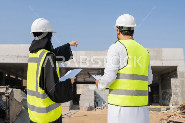 Follow-up and supervision of engineering and construction projects, studying the plan and the project on the ground, an engineering profession and job, an Arab Gulf Emirati engineer wearing a kandura, jacket and protective helmet working with his colleague in the engineering sector in the United Arab Emirates, monitoring and managing construction operations.