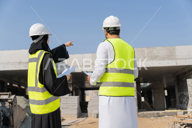 Engineering and construction project management, studying the plan and the project on the ground, an engineering profession and job, an Arab Gulf Emirati engineer wearing a kandura, jacket and protective helmet working with his colleague in the engineering sector in the United Arab Emirates, monitoring and following up on construction operations.