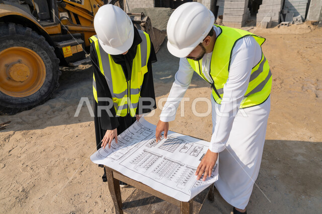 المتابعة والاشراف على المشاريع الهندسية والانشاءات، دراسة المخطط والمشروع على ارض الواقع، مهنة ووظيفة هندسية، مهندس عربي خليجي اماراتي يرتدي الكندورة والسترة وخوذة الحماية يعمل مع زميلته في القطاع الهندسي في الامارات العربية المتحدة، مراقبة وادارة عمليات البناء