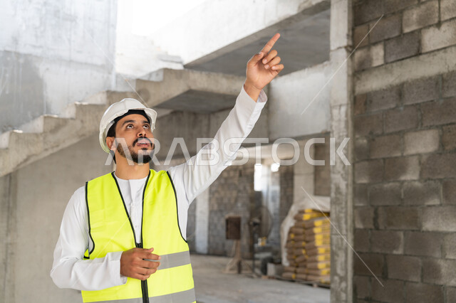 الاشارة الى شيء ما، مهنة هندسية شبابية، مهندس عربي خليجي اماراتي يرتدي الكندورة والسترة وخوذة الحماية يعمل في القطاع الهندسي في الامارات العربية المتحدة، الاشرف على عمليات البناء، مفهوم ادارة المشاريع الهندسية والانشاءات، دراسة المخطط والمشروع على ارض الواقع