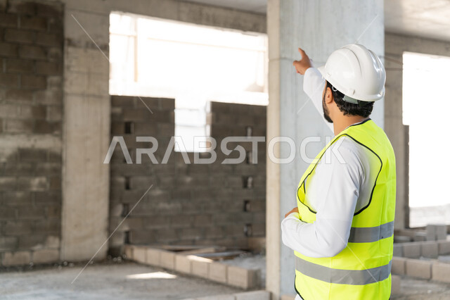 Pointing to something, a youth engineering profession, an Arab Gulf Emirati engineer wearing a kandura, jacket and protective helmet working in the engineering sector in the United Arab Emirates, supervising construction operations, the concept of engineering and construction project management, studying the plan and the project on the ground