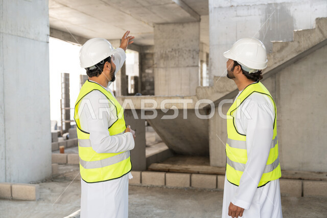 The concept of engineering and construction project management, studying the plan and the project on the ground, a youth engineering profession and job, two Arab Gulf Emirati engineers wearing kanduras, jackets and protective helmets working in the engineering sector in the Emirates, monitoring and following up on construction operations.