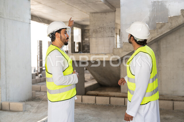 Monitoring and following up on construction operations, the concept of engineering and construction project management, studying the plan and project on the ground, a youth engineering profession and job, two Arab Gulf Emirati engineers wearing kanduras, jackets and protective helmets working in the engineering sector in the Emirates.