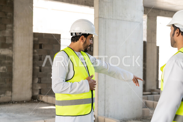 The concept of engineering and construction project management, studying the plan and the project on the ground, a youth engineering profession and job, two Arab Gulf Emirati engineers wearing kanduras, jackets and protective helmets working in the engineering sector in the Emirates, monitoring and following up on construction operations.