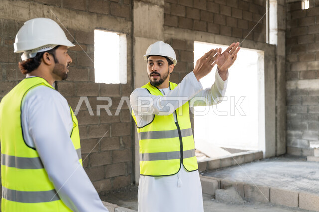 مراقبة ومتابعة عمليات البناء، مفهوم ادارة المشاريع الهندسية والانشاءات، دراسة المخطط والمشروع على ارض الواقع، مهنة ووظيفة هندسية شبابية، مهندسان عربيان خليجيان اماراتيان يرتديان الكندورة والسترة وخوذة الحماية يعملان في القطاع الهندسي في الامارات