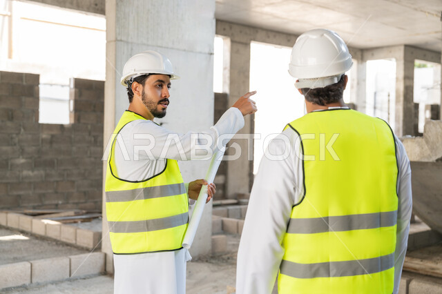 مراقبة ومتابعة عمليات البناء، مفهوم ادارة المشاريع الهندسية والانشاءات، دراسة المخطط والمشروع على ارض الواقع، مهنة ووظيفة هندسية شبابية، مهندسان عربيان خليجيان اماراتيان يرتديان الكندورة والسترة وخوذة الحماية يعملان في القطاع الهندسي في الامارات