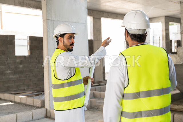 The concept of engineering and construction project management, studying the plan and the project on the ground, a youth engineering profession and job, two Arab Gulf Emirati engineers wearing kanduras, jackets and protective helmets working in the engineering sector in the Emirates, monitoring and following up on construction operations.