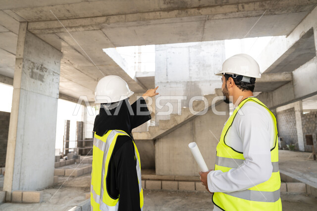 Engineering and construction project management, studying the plan and the project on the ground, an engineering profession and job, an Arab Gulf Emirati engineer wearing a kandura, jacket and protective helmet working with his colleague in the engineering sector in the United Arab Emirates, monitoring and following up on construction operations.