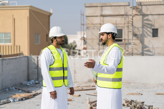 مفهوم ادارة المشاريع الهندسية والانشاءات، دراسة المخطط والمشروع على ارض الواقع، مهنة ووظيفة هندسية شبابية، مهندسان عربيان خليجيان اماراتيان يرتديان الكندورة والسترة وخوذة الحماية يعملان في القطاع الهندسي في الامارات، مراقبة ومتابعة عمليات البناء