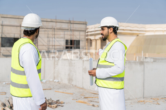 مفهوم ادارة المشاريع الهندسية والانشاءات، دراسة المخطط والمشروع على ارض الواقع، مهنة ووظيفة هندسية شبابية، مهندسان عربيان خليجيان اماراتيان يرتديان الكندورة والسترة وخوذة الحماية يعملان في القطاع الهندسي في الامارات، مراقبة ومتابعة عمليات البناء