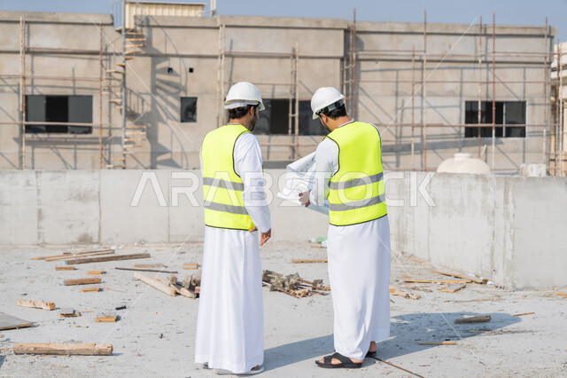 مفهوم ادارة المشاريع الهندسية والانشاءات، دراسة المخطط والمشروع على ارض الواقع، مهنة ووظيفة هندسية شبابية، مهندسان عربيان خليجيان اماراتيان يرتديان الكندورة والسترة وخوذة الحماية يعملان في القطاع الهندسي في الامارات، مراقبة ومتابعة عمليات البناء