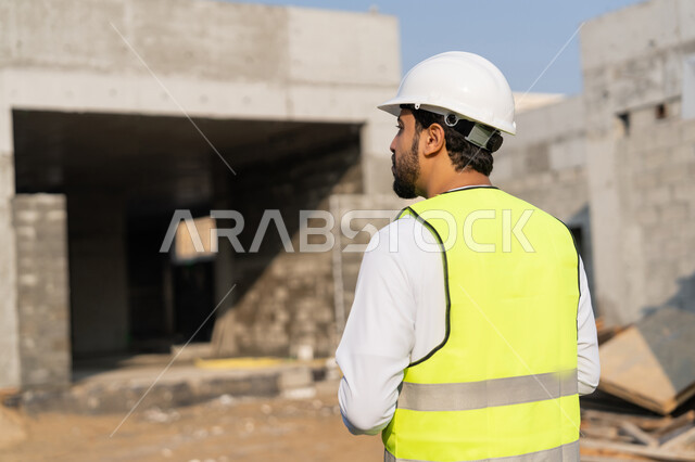 The concept of engineering and construction project management, studying the plan and the project on the ground, a youth engineering profession and job, an Arab Gulf Emirati engineer wearing a kandura, jacket and protective helmet, working in the engineering sector in the United Arab Emirates, supervising construction operations.