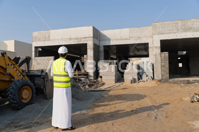 مفهوم ادارة المشاريع الهندسية والانشاءات، دراسة المخطط والمشروع على ارض الواقع، مهنة ووظيفة هندسية شبابية، مهندس عربي خليجي اماراتي يرتدي الكندورة والسترة وخوذة الحماية يعمل في القطاع الهندسي في الامارات العربية المتحدة، الاشرف على عمليات البناء