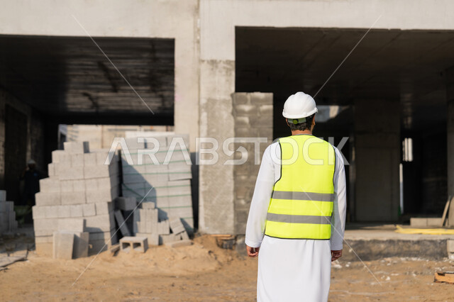 The concept of engineering and construction project management, studying the plan and the project on the ground, a youth engineering profession and job, an Arab Gulf Emirati engineer wearing a kandura, jacket and protective helmet, working in the engineering sector in the United Arab Emirates, supervising construction operations.