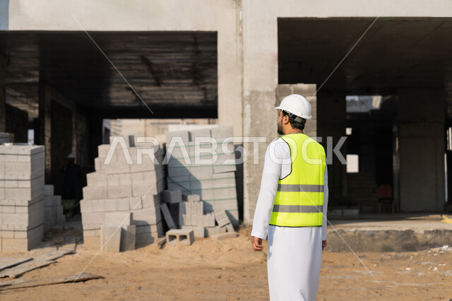 مهنة ووظيفة هندسية شبابية، مهندس عربي خليجي اماراتي يرتدي الكندورة والسترة وخوذة الحماية يعمل في القطاع الهندسي في الامارات العربية المتحدة، الاشرف على عمليات البناء، مفهوم ادارة المشاريع الهندسية والانشاءات، دراسة المخطط والمشروع على ارض الواقع