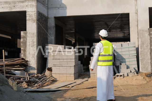 A youth engineering profession and job, an Arab Gulf Emirati engineer wearing a kandura, jacket and protective helmet, working in the engineering sector in the United Arab Emirates, supervising construction operations, the concept of engineering and construction project management, studying the plan and the project on the ground