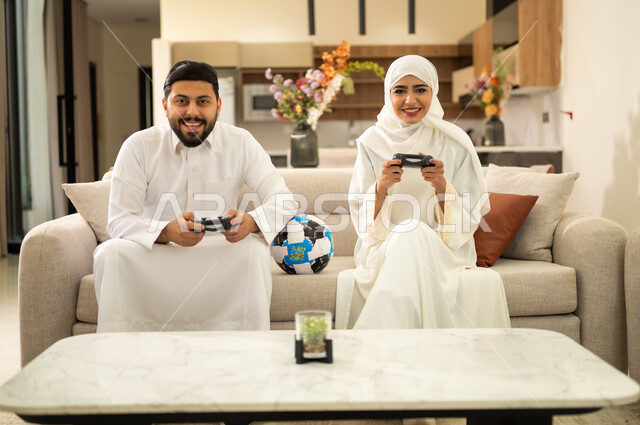 التفاعل والاندماج مع العاب الفيديو ،الاستمتاع بالتقنيات والتكنولوجيا الحديثة ، زوجان عربيان خليجيان سعوديان يمسكان عصا التحكم وبجانبهما كرة القدم، قضاء اوقات ممتعة مع العائلة ، فعاليات عطلة نهاية الأسبوع  .