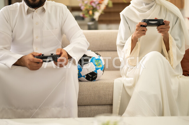 مفهوم المتعة والتسلية ،  استخدام التكنولوجيا الحديثة، صورة مقربة ليد زوجان عربيان خليجيان سعوديان يمسكان عصا التحكم وبجانبهما كرة القدم، قضاء اوقات ممتعة مع العائلة ، التفاعل والاندماج مع الالعاب الالكترونية .