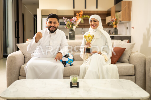 Gestures of joy and enthusiasm for victory, spending enjoyable leisure time with the wife, interacting with the national team, matches and sports, happiness for winning the cup, an Arab Gulf Saudi couple watching and following football with expressions of victory.