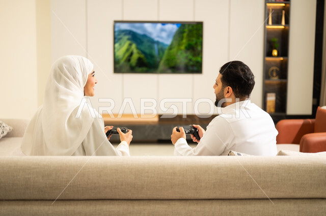 لعب العاب الفيديو الترفيهية ، قضاء أوقات الفراغ مع العائلة ، استخدام التكنولوجيا الحديثة، صورة من الخلف لزوجان عربيان خليجيان سعوديان يمسكان عصا التحكم، إيماءات الفوز والخسارة فى اللعبة ، التفاعل والاندماج مع الالعاب الالكترونية   .