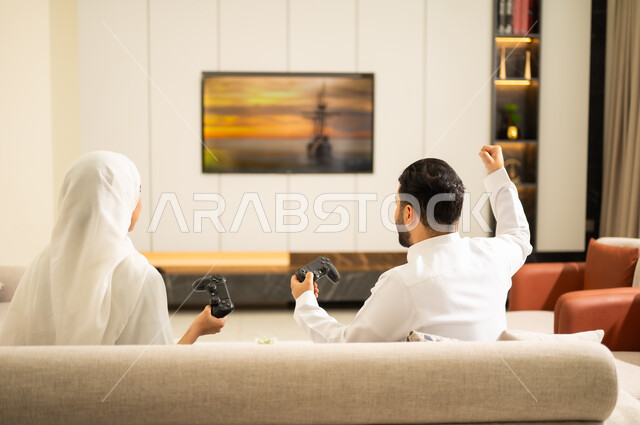 اجهزة الكترونية وتقنيات حديثة ، فعاليات عطلة نهاية الأسبوع  ، العالم الرقمي الترفيهي ، ايماءات الفوز والخسارة ، المتعة والتسلية بالعاب الفيديو ، صورة من الخلف لزوجان عربيان خليجيان سعوديان يمسكان عصا التحكم ، يقومان بلعب الالعاب الالكترونية ، ممارسة الأنشطة الترفيهية وقضاء أوقات الفراغ  .