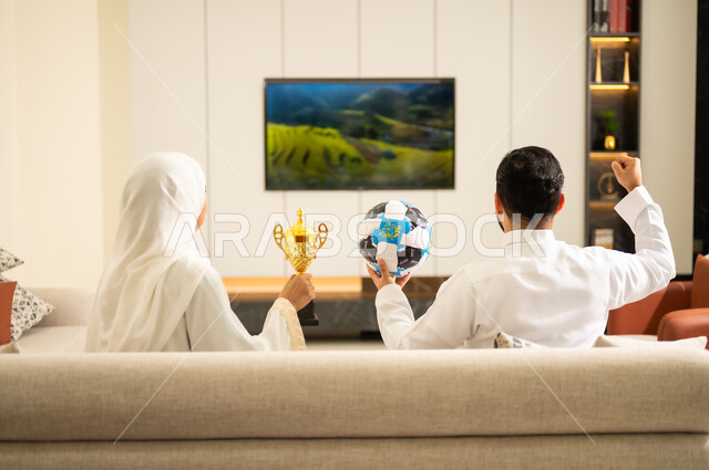 الانتصار بتحقيق الاهداف ، ايماءات الشغف والتشجيع للفريق، الحماس والتفاعل مع المنتخب ،المباريات والألعاب الرياضية,  قضاء اوقات الفراغ الممتعة مع الزوجة ، صورة من الخلف لزوجان عربيان خليجيان سعوديان يجلسان على أريكة مريحة يمسك الزوج كرة بيده وتحمل زوجته كأس ، يقومان بمشاهدة الفريق المفضل .