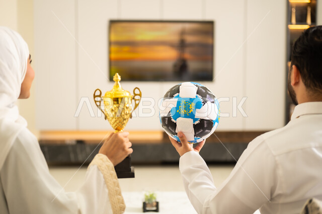 الحماس بالفوز بالكأس ، قضاء اوقات الفراغ الممتعة مع الزوجة  ، ايماءات الشغف والتشجيع للفريق، الحماس والتفاعل مع المنتخب ،المباريات والألعاب الرياضية، صورة مقربة ليد زوجان عربيان خليجيان سعوديان يمسك الزوج كرة بيده وتحمل زوجته كأس ، يقومان بمشاهدة الفريق المفضل بايماءات الانتصار.
